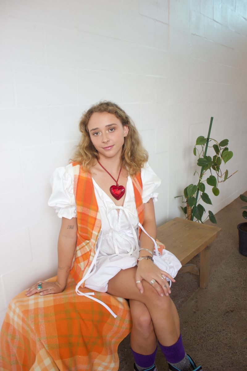 Woman sits on a bench wearing a large red necklace, white milkmaid dress with orange accents