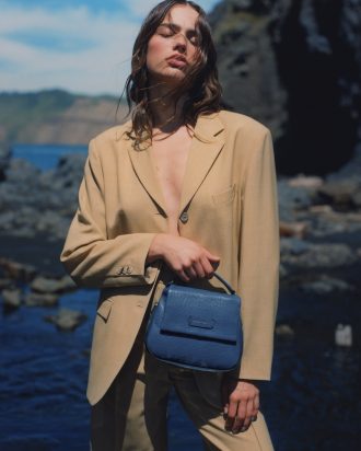 Woman is posing with her eyes closed, clutching a blue leather bag. She is wearing a camel coloured blazer and pant set. Photo features deep blue ocean backdrop