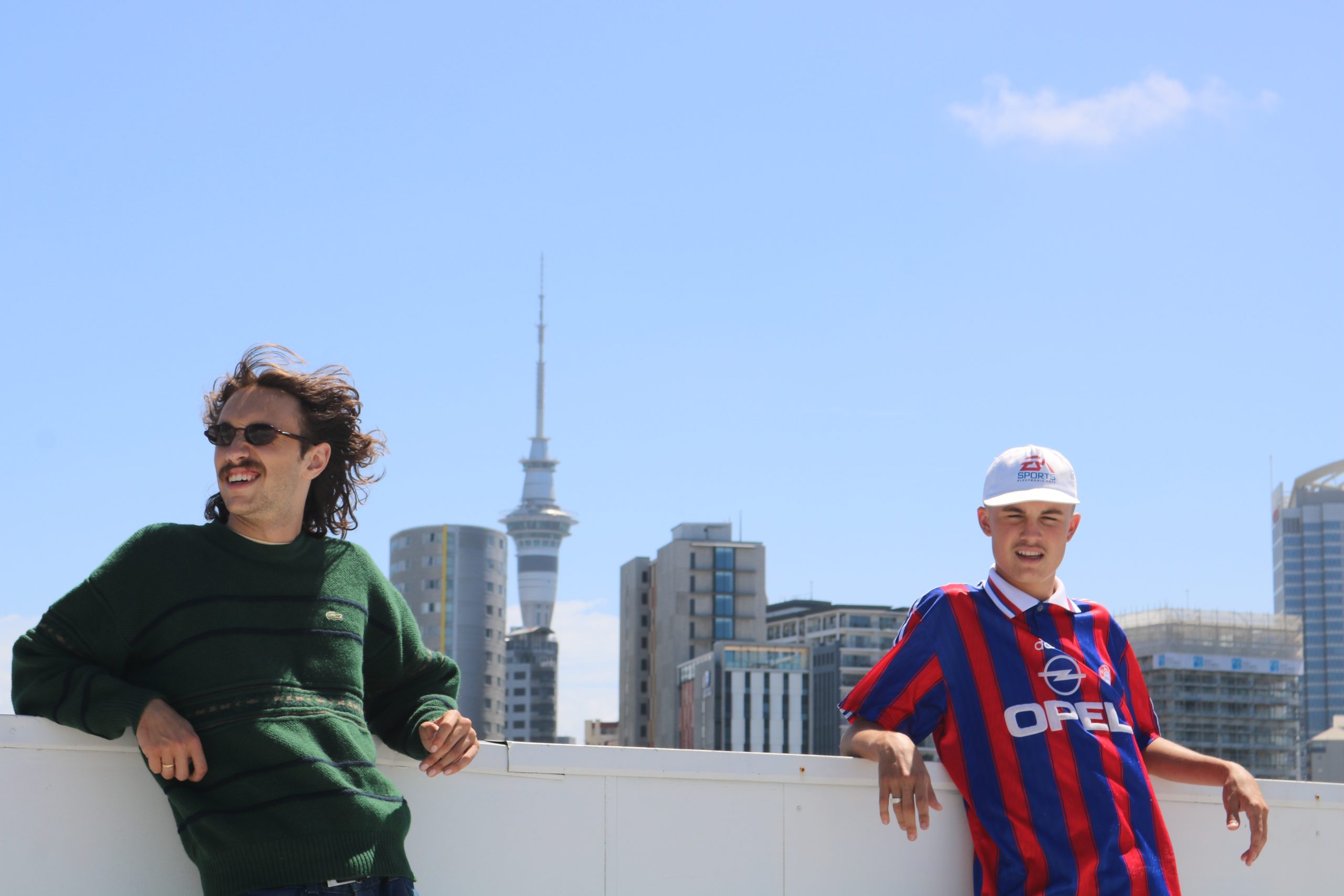 Two men are pictured on a rooftop carpark looking towards the left. The Auckland skytower can be seen in the horizon.