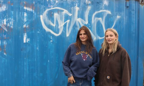 Two women are pictured behind a blue painted background with white grafitti. The woman on the left is wearing a navy Guess sweatshirt while the woman on the right is wearing a brown zip-up hoodie