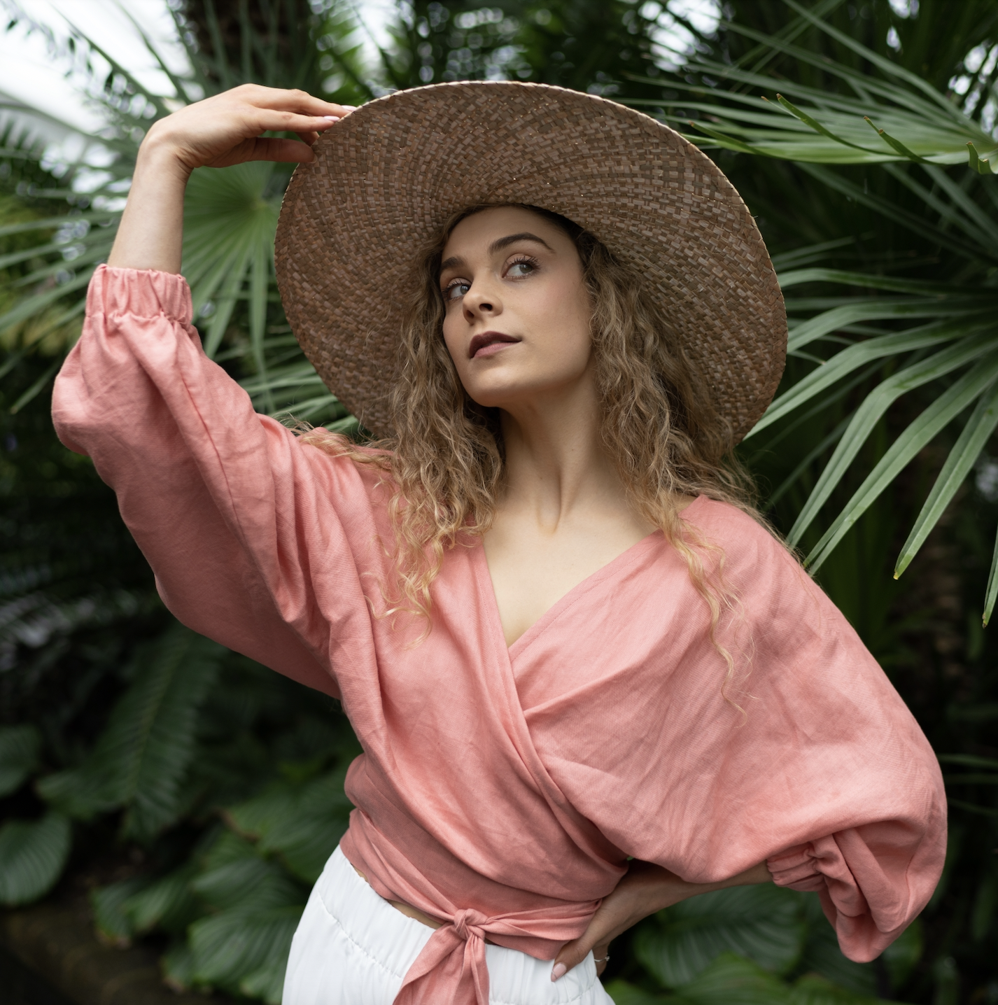 Woman standing in front of palm leaves in large sun hat and peach Danni Rose wrap top