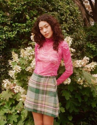 Woman posing in a garden, wearing a pink long sleeve top and a green plaid a-line skirt