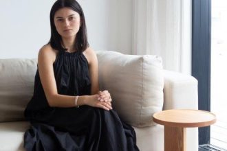 New Zealand designer Poppy Voon is pictured sitting on a grey couch in a black sleeveless dress