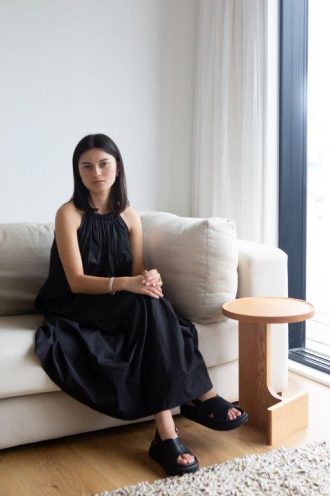 New Zealand designer Poppy Voon is pictured sitting on a grey couch in a black sleeveless dress