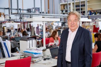 Landscape image of man in black and white casual suit leaning on a desk in a fashion work space. In the background, multiple people are working on designs