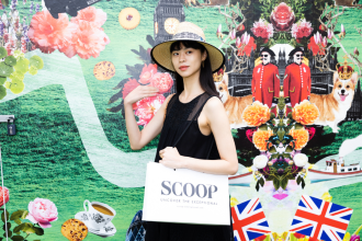 Image of woman standing in front of green feature wall, in a black dress and white hat. She is holding a white bag with the scoop logo.