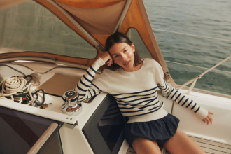 Woman in white jumper with blue stripes lounges on sailing boat, looking into the camera