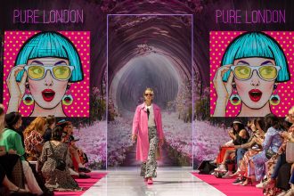 Woman in pink coat and patterned trousers walks down pink catwalk. An animated image of a woman with a blue bob cut is projected on two screens behind her.