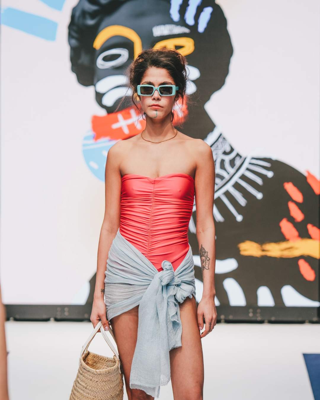 A woman walking down the runway. She is wearing blue glasses, a pink corset and a wrap around skirt.