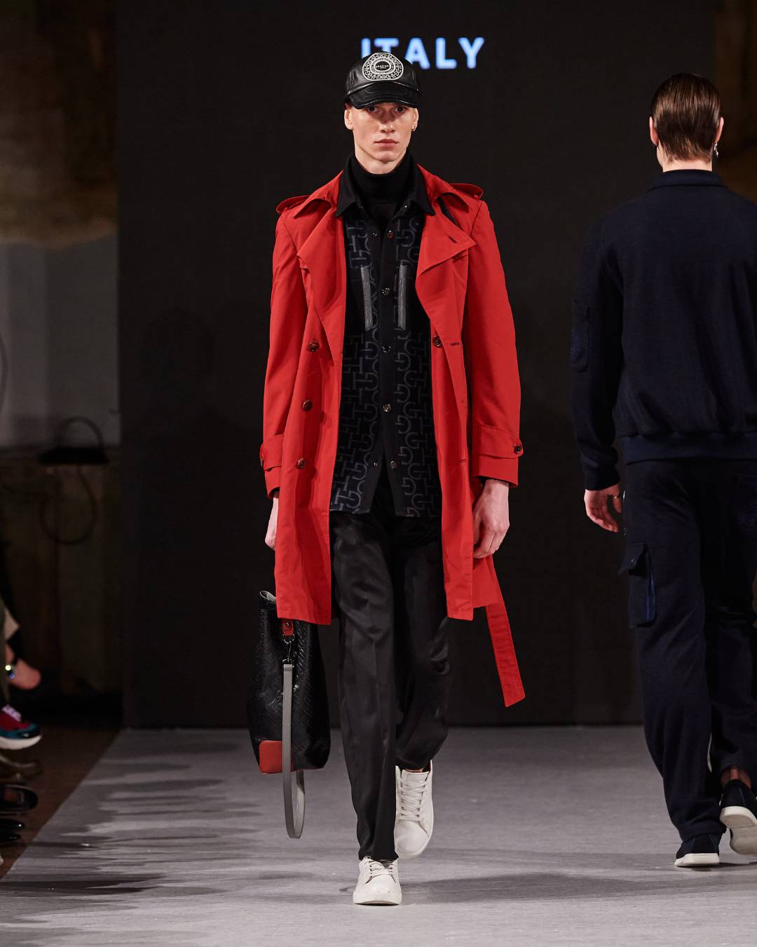 A male model walking down the runway with a red trenchcoat on and a black cap.